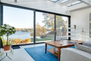 Sala integrada com grandes portas e janelas de esquadrias de alto padrão em fachada residencial moderna, com parede branca, piso em porcelanato e vista para o lago.